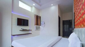 A modern hotel room with a large bed, a flat-screen TV mounted on the wall, a small shelf, and a window with curtains. - Hotel Vishnu Residency Inn (Amritsar)
