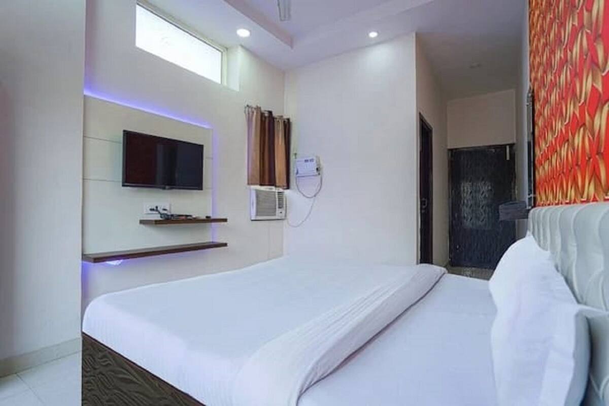 Quarto de hotel moderno com uma cama grande, uma televisão de tela plana na parede, uma pequena estante e uma janela com cortinas.