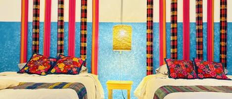 Habitación de hotel con dos camas, cortinas coloridas, un taburete pequeño y un tapiz decorativo.