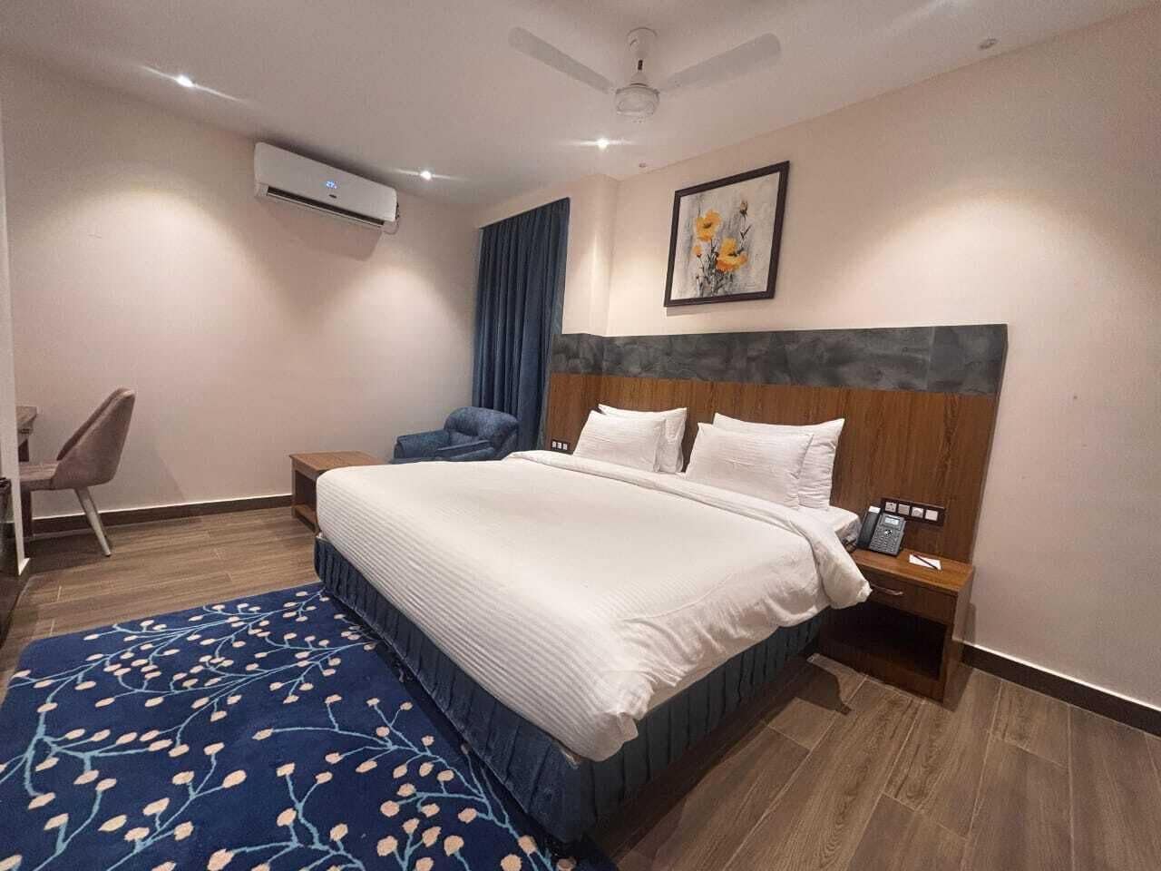 A hotel room with a large bed, a bedside table with a phone, a chair, a desk, and an air conditioning unit on the wall.