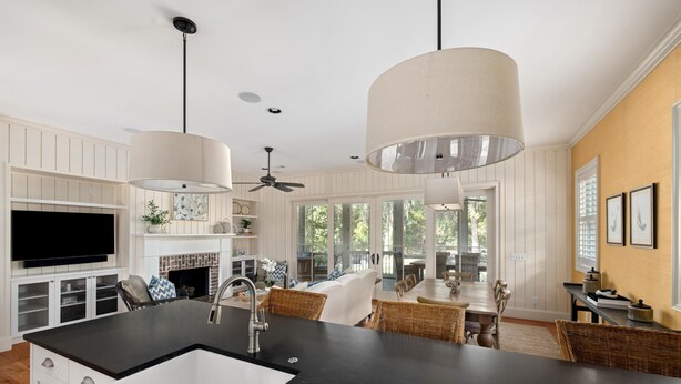 A modern kitchen with a black countertop, white cabinets, and a large island with a sink. There is a dining area with wicker chairs and a table, and a ceiling fan. - Wilson Cottages, Walk to Tennis & Pickleball, and Restaurants (Bluffton)