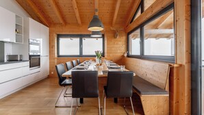 Una cocina moderna con armarios blancos y electrodomésticos de acero inoxidable, un comedor con mesa y sillas de madera, y ventanas amplias que ofrecen luz natural.