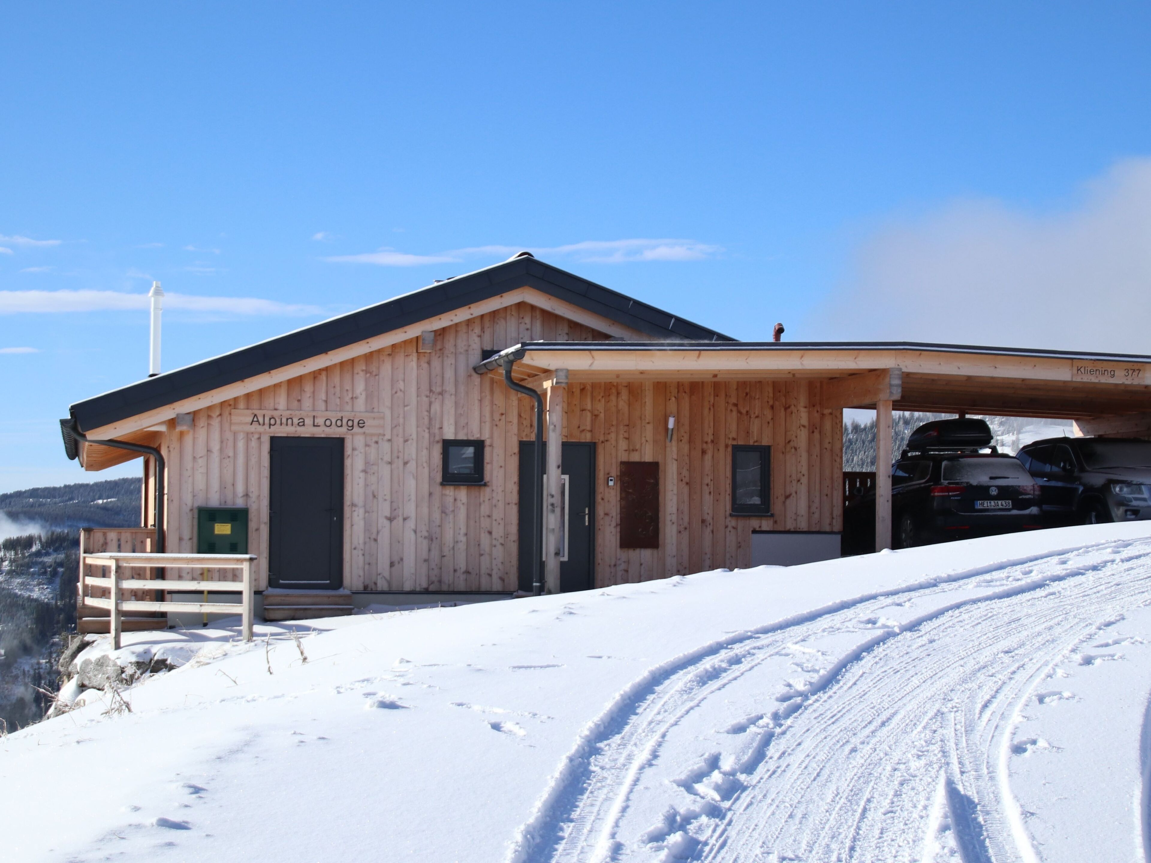 Una cabaña de madera llamada 'Alpina Lodge', con zona de estacionamiento techada y una ladera nevada al frente.