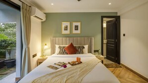 A hotel room with a bed, bedside table, lamp, and a view of the outdoors. - Luxury 3BR Duplex With Pool in Goa s Beverly Hills (Assagao)
