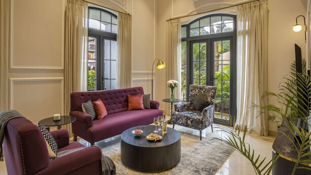 A spacious living room with a red sofa, a patterned armchair, a round coffee table, and large windows with curtains. - Luxury 3BR Duplex With Pool in Goa s Beverly Hills (Assagao)