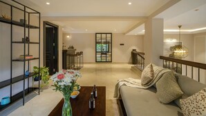 A spacious living room with a sofa, coffee table, and decorative items. There is a bookshelf, a dining area, and a view through a glass door. - Luxury 3BR Duplex With Pool in Goa s Beverly Hills (Assagao)
