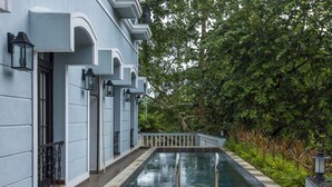 A pool area with a long rectangular pool, surrounded by a low white fence, and adjacent to a building with a light blue facade and decorative arches. - Luxury 3BR Duplex With Pool in Goa s Beverly Hills (Assagao)