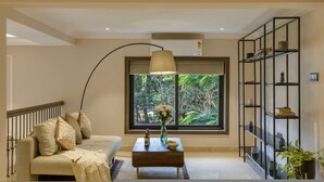 A modern living room with a sofa, coffee table, and a large window with a view of greenery. - Luxury 3BR Duplex With Pool in Goa s Beverly Hills (Assagao)