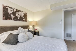 Una cama bien hecha con almohadas, un mapa del mundo en la pared, una lámpara y una puerta a otra habitación.