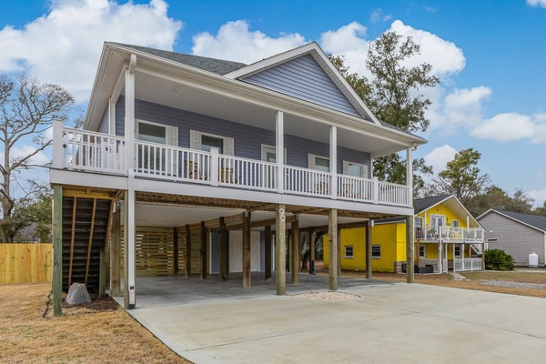 Exterior view of the home, stairs leading to the second floor, a balcony with seating, and ample parking both under the home and in the driveway.