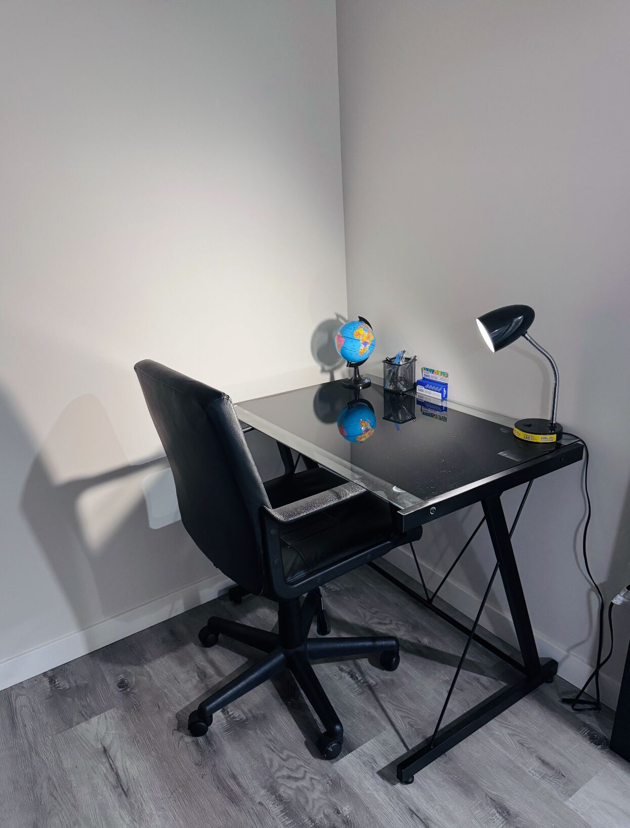 A small office space with a black office chair, a glass desk, a globe, a lamp, and a pen holder.