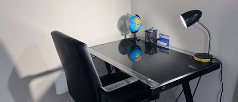 A small office space with a black office chair, a glass desk, a globe, a lamp, and a pen holder.