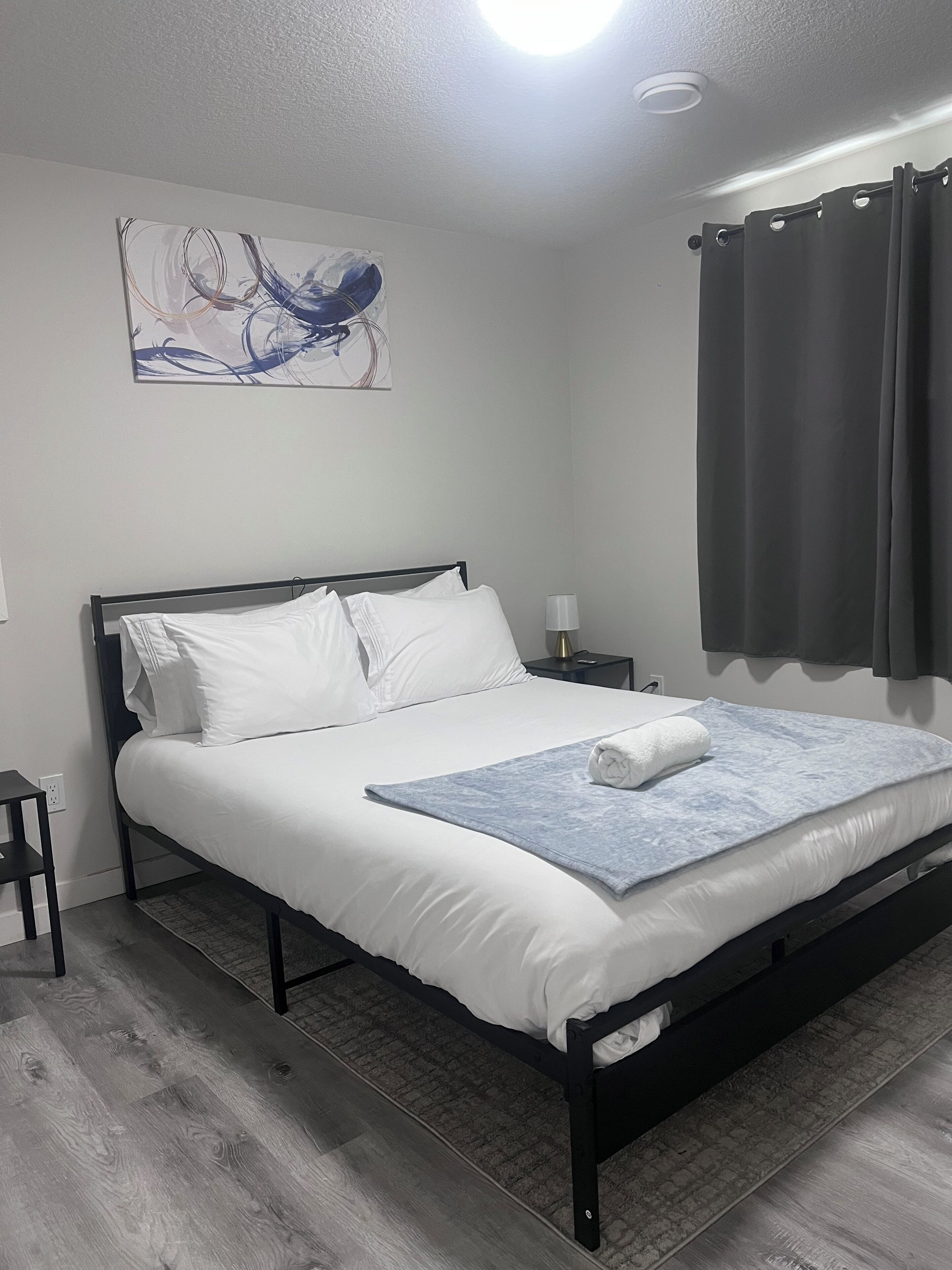 Een net opgemaakt bed met witte lakens, een blauwe plaid en een opgerolde handdoek. Er staat een nachtkastje met een lamp naast het bed, en een schilderij hangt aan de muur erboven.