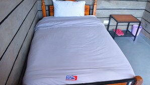 Un letto singolo con struttura e testiera in legno, un materasso bianco e un comodino in legno con un cesto e ciabatte.