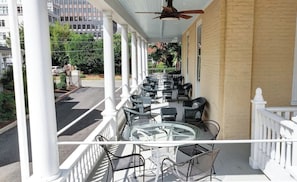 A covered outdoor seating area with chairs and tables, white railings, and a view of the street and buildings.