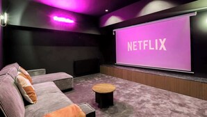 Una sala de cine en casa moderna con una pantalla grande mostrando el logotipo de Netflix, un área de estar cómoda con un sofá y una mesita redonda, y una pared oscura con paneles de madera.