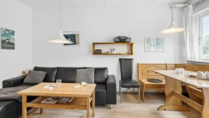 Un espacio de estar moderno con sofá, una mesa de centro de madera, una mesa de comedor y un banco. Hay dos lámparas colgantes, una estantería con una radio y obras de arte enmarcadas en las paredes.