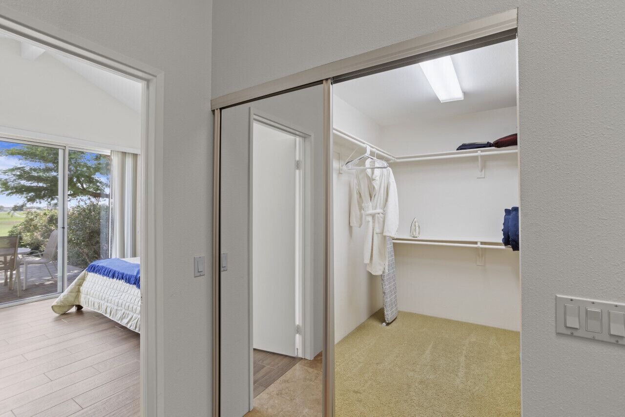 Une chambre avec une grande porte-fenêtre coulissante donnant sur un balcon, un lit recouvert d’une courtepointe bleue et blanche, une robe blanche suspendue à un porte-vêtements et une étagère blanche sur laquelle se trouve un récipient bleu.