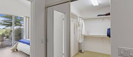 Une chambre avec une grande porte-fenêtre coulissante donnant sur un balcon, un lit recouvert d’une courtepointe bleue et blanche, une robe blanche suspendue à un porte-vêtements et une étagère blanche sur laquelle se trouve un récipient bleu.
