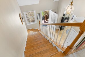 Una escalera de madera con barandillas blancas, vistas al exterior a través de una puerta, y una estantería llena de libros.