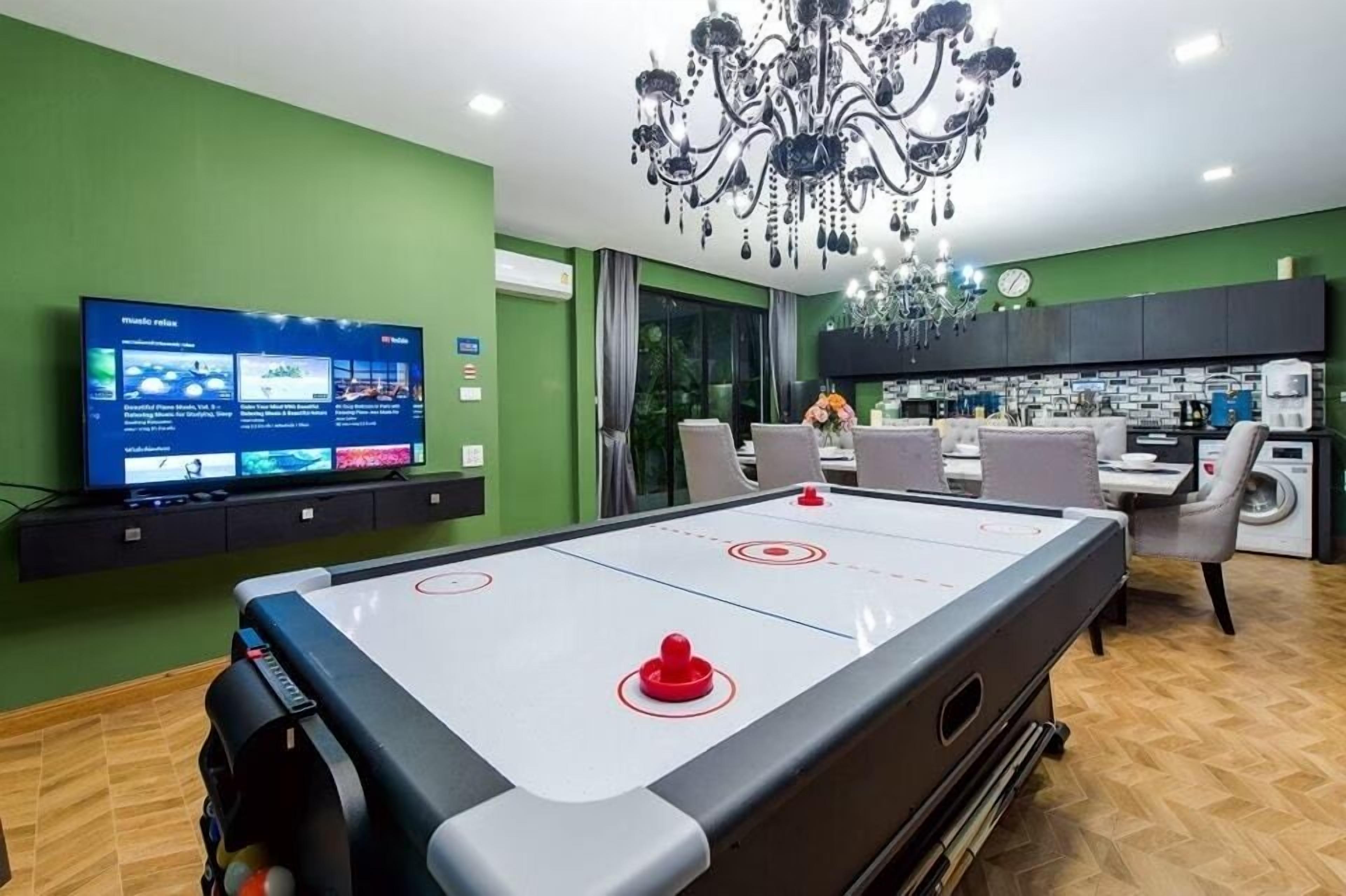 A pool table with a TV displaying a sports channel, a chandelier, a dining area, and a kitchen in the background.