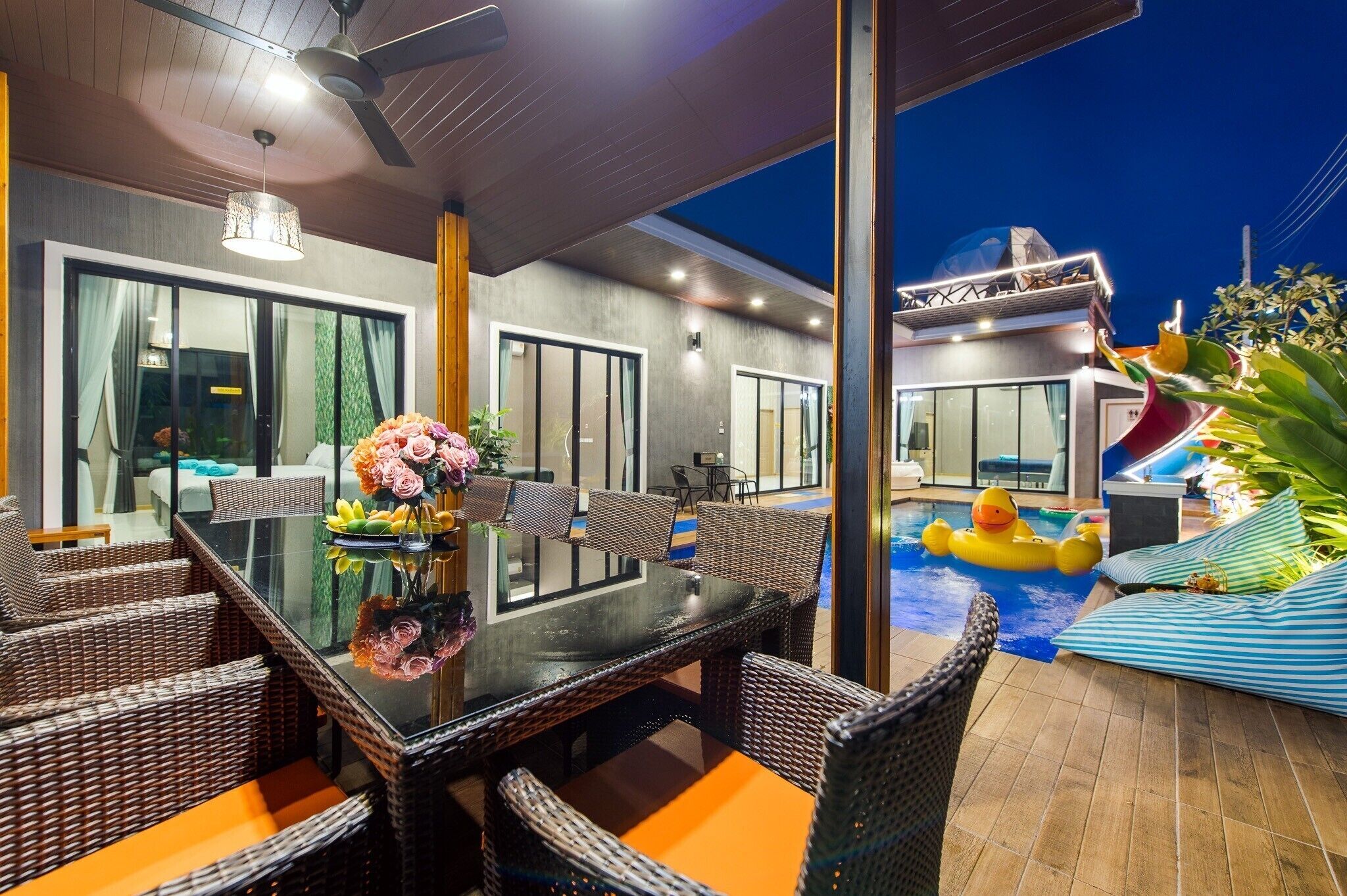 Un espacio moderno para comer al aire libre con mobiliario de mimbre y una mesa de vidrio, con vista a una piscina que tiene un tobogán y un pato inflable.