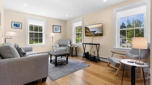A living room with a sofa, armchairs, a coffee table, and a TV. There are large windows with views of greenery and a landscape. - Puffins Perch, romantic waterfront Maine cottage (Saint George)