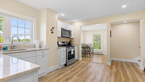 A kitchen with white cabinets, a microwave, an oven, and a dining area with a table and chairs. - Puffins Perch, romantic waterfront Maine cottage (Saint George)