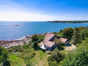 A coastal house with a sloped roof, surrounded by greenery and a view of the sea.