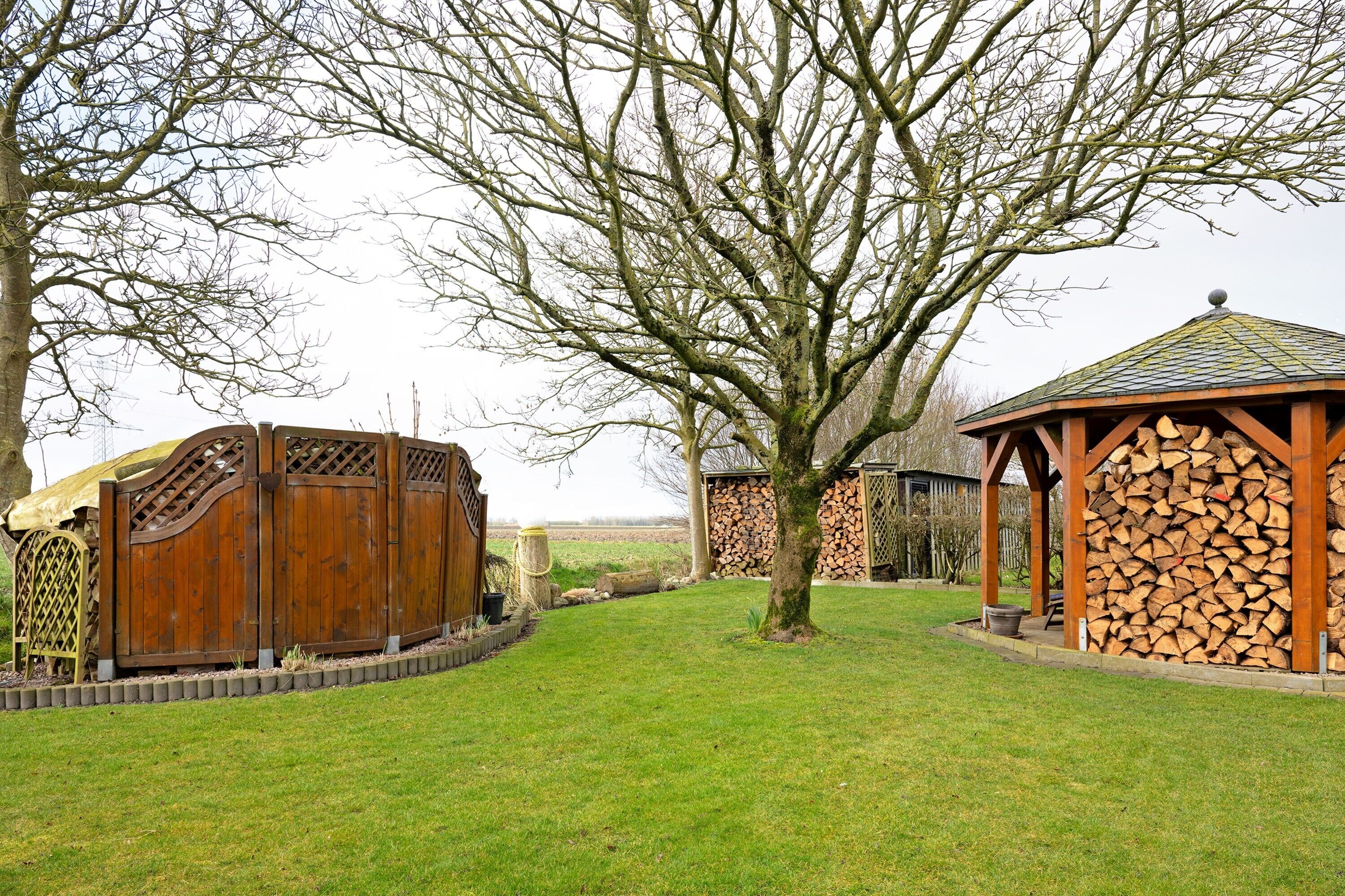 A wooden gazebo, a tree, and a stack of firewood in a grassy yard.