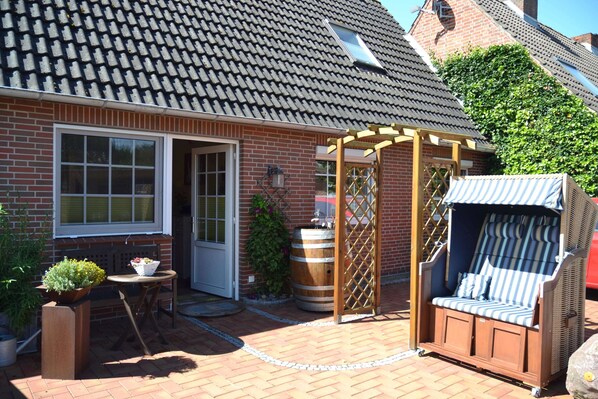 Una casa de ladrillo con techo de tejas, una pérgola de madera, una silla de mimbre, un barril y una planta en maceta.