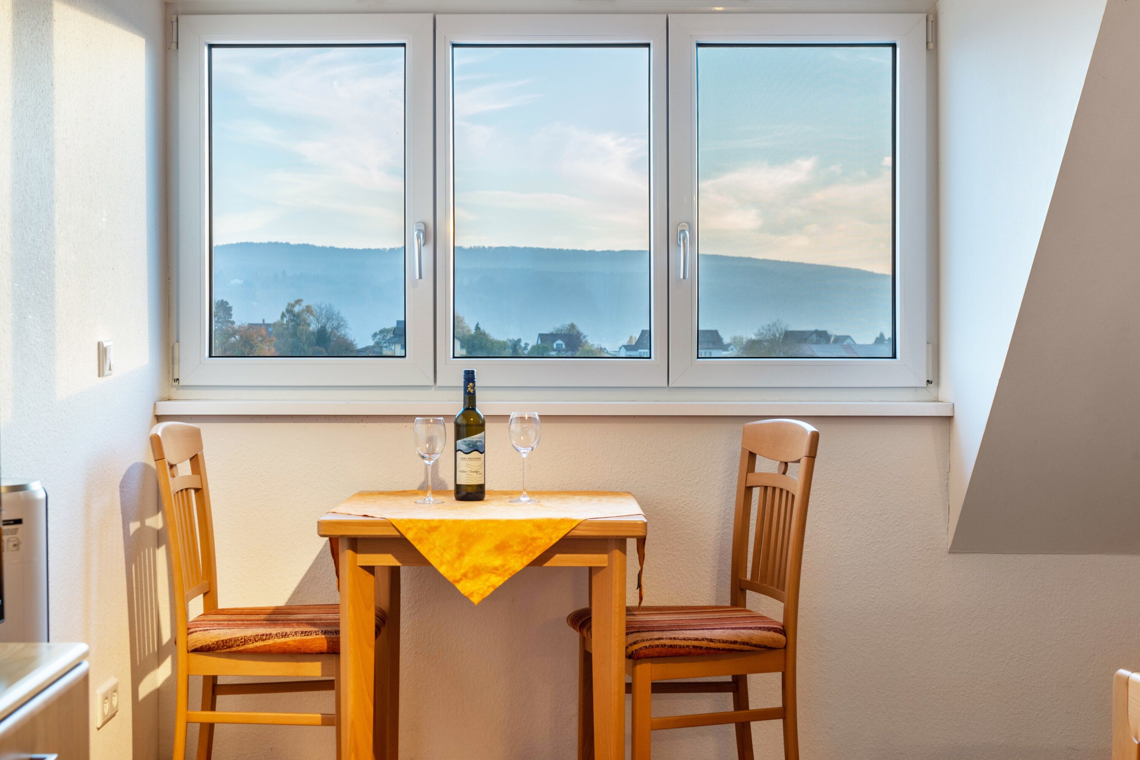 A room with a table set for two, a bottle of wine, and two glasses. There is a window with a view of a landscape.