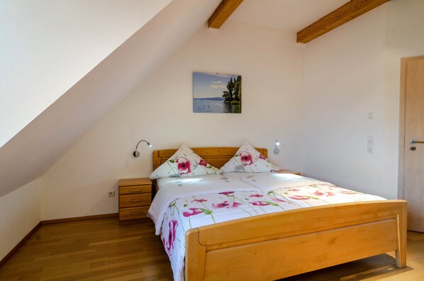 A bedroom with a wooden bed, a nightstand, a dresser, a painting of a lake, and a sloped ceiling.