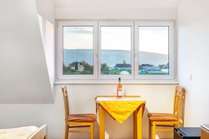 Quarto com uma mesa e duas cadeiras, janela com vista para edifícios e colinas, e uma garrafa sobre a mesa.