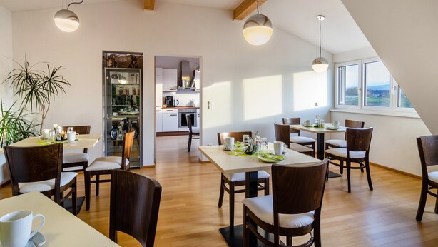 A dining area with wooden tables and chairs, a kitchen in the background, and large windows with a view. - Room 'Zimmer 2' with Shared Garden and Wi-Fi (Reichenau)