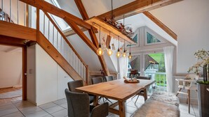 Un comedor amplio con una mesa de madera, sillas grises y un banco de piel. Se aprecia una escalera con vigas de madera y un tragaluz al fondo.