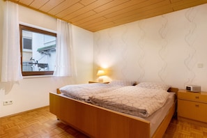 A bedroom with a wooden bed, a bedside table with a lamp, a nightstand, and a window with curtains.