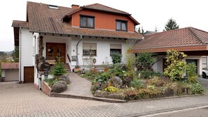 A two-story house with a tiled roof, a front porch, and a garden with various plants and a bench.