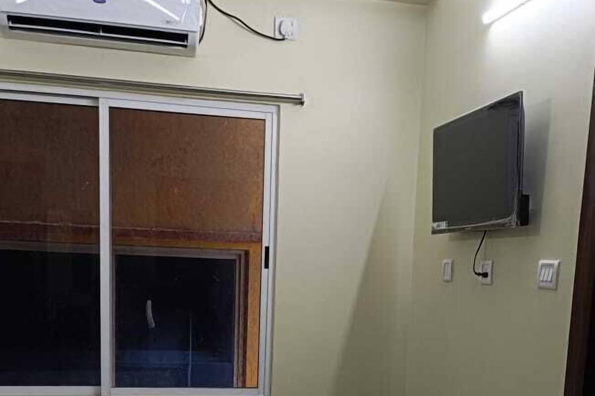 A bedroom with a bed, a small table, an air conditioning unit, a TV, and a window with blinds.