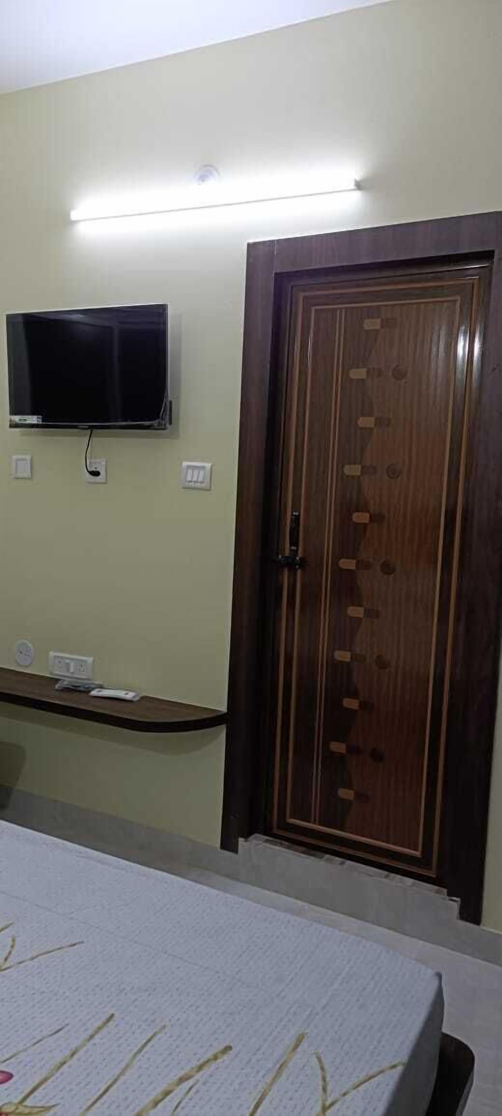 A room with a bed, a TV mounted on the wall, a wooden wardrobe, and a bedside table with a power outlet.