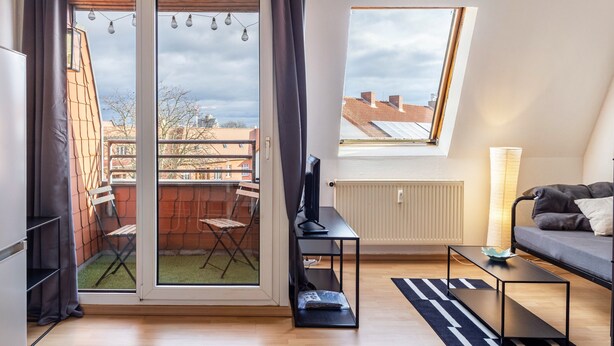 Un appartement moderne doté d’une porte-fenêtre coulissante donnant sur un balcon, d’un téléviseur posé sur un meuble, d’un canapé et d’une fenêtre offrant une vue sur les immeubles.