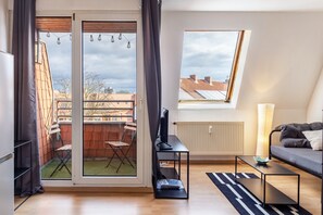 Un apartamento moderno con puerta de vidrio corredera a un balcón, un televisor sobre un soporte, un sofá y una ventana con vista a edificios.