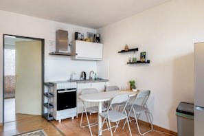 A compact kitchen with a stove, oven, and microwave, a dining table with chairs, a small shelf with books and a plant, and a trash bin.