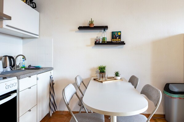 A compact kitchen with a white countertop, a stove, a sink, and a dining table with chairs. There are two shelves with plants and a book.