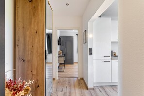 A modern interior with a wooden accent wall, a kitchen with white cabinets, and a hallway leading to a room with a dark-colored door.