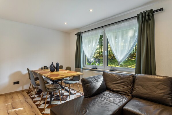 A modern living room with a leather sofa, a dining table with chairs, and a large window with curtains.