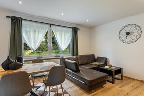 A modern living room with a sofa, dining table, chairs, and a view through large windows.