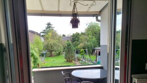 Un balcon avec une table et des chaises, une plante suspendue, et une vue sur un jardin et les maisons voisines.