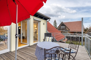 Um terraço com mesa e cadeiras, um guarda-sol vermelho e um edifício com telhado vermelho.