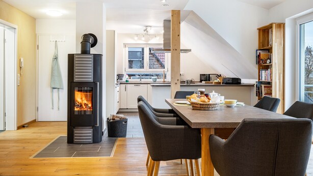 Una cocina moderna con estufa de leña, una mesa de comedor con sillas y una escalera que conduce a un nivel superior.